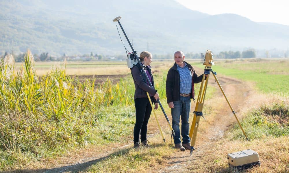Surveyors conducting topographic surveys in a rural field using surveying equipment with hills in the background.
