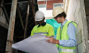 Two hard-hat workers in safety vests reviewing large building plans at a construction site